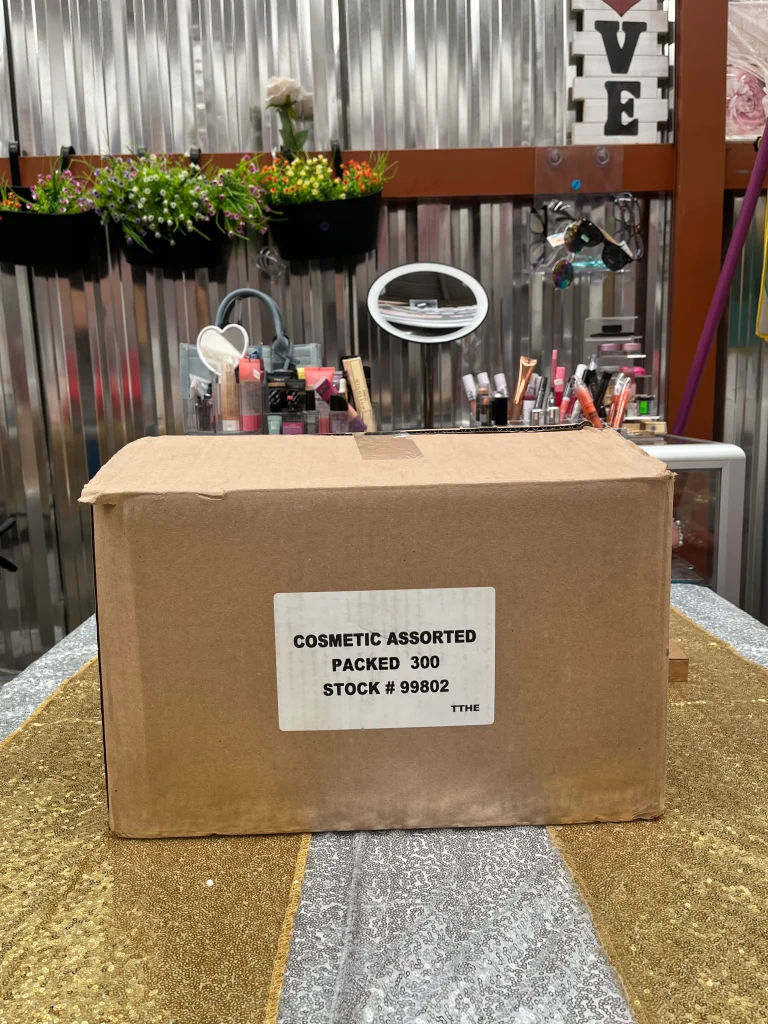 Imagen de una caja de cartón con etiqueta “Cosmetic Assorted Packed 300 Stock #99802”, colocada sobre una mesa decorada, en un entorno con productos de maquillaje y elementos decorativos al fondo.